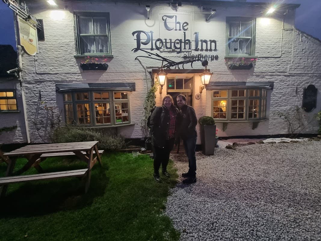 Two women stand outside The Plough Inn, a charming pub with a gravel path and lanterns, surrounded by greenery.