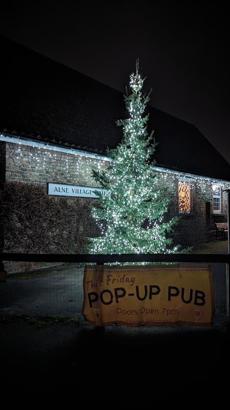 Alne Village Hall illuminated at night with a decorated Christmas tree and a banner for a pop-up pub event.