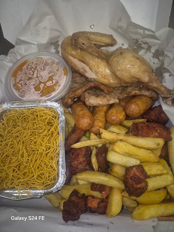 Takeaway box featuring fried chicken, sausages, chips, and noodles, with a container of sauce on the side.