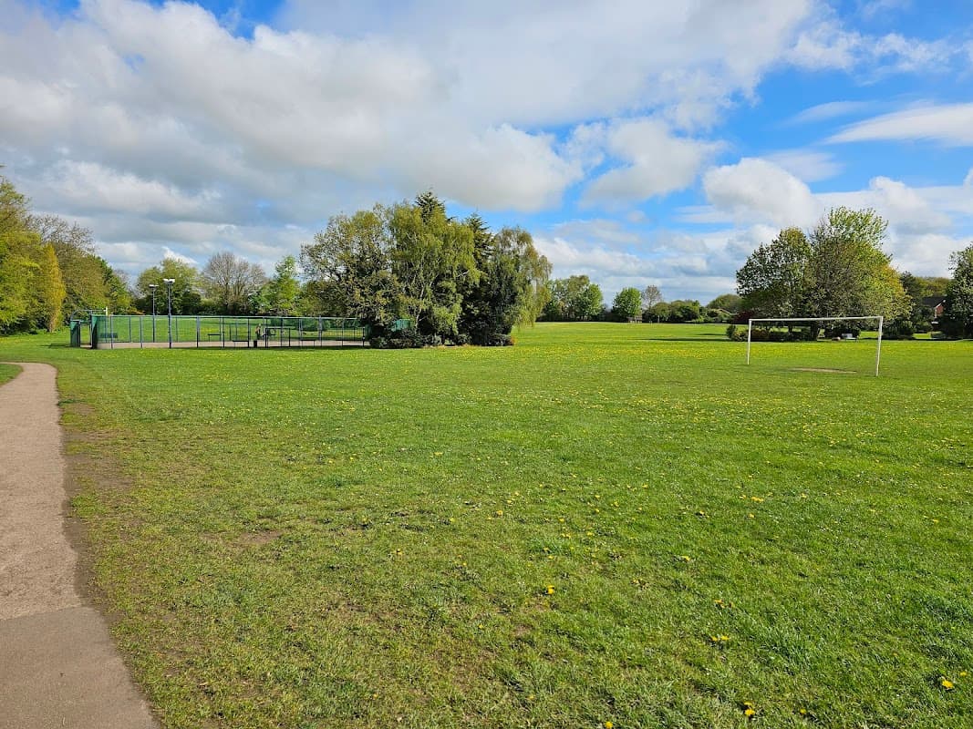 Spacious green park with a football goal, playground area, and scattered trees under a partly cloudy sky.