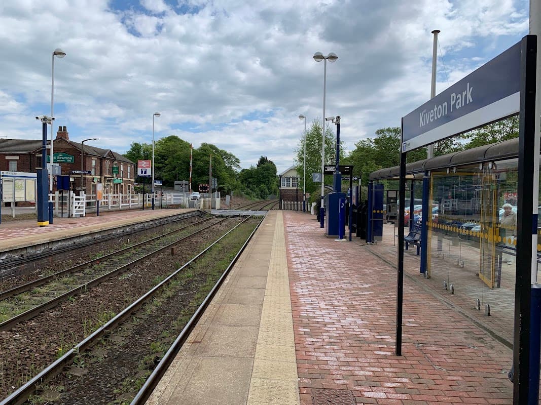Kiveton Park bus stop with brick pathway, railway tracks, and green trees under a cloudy sky.