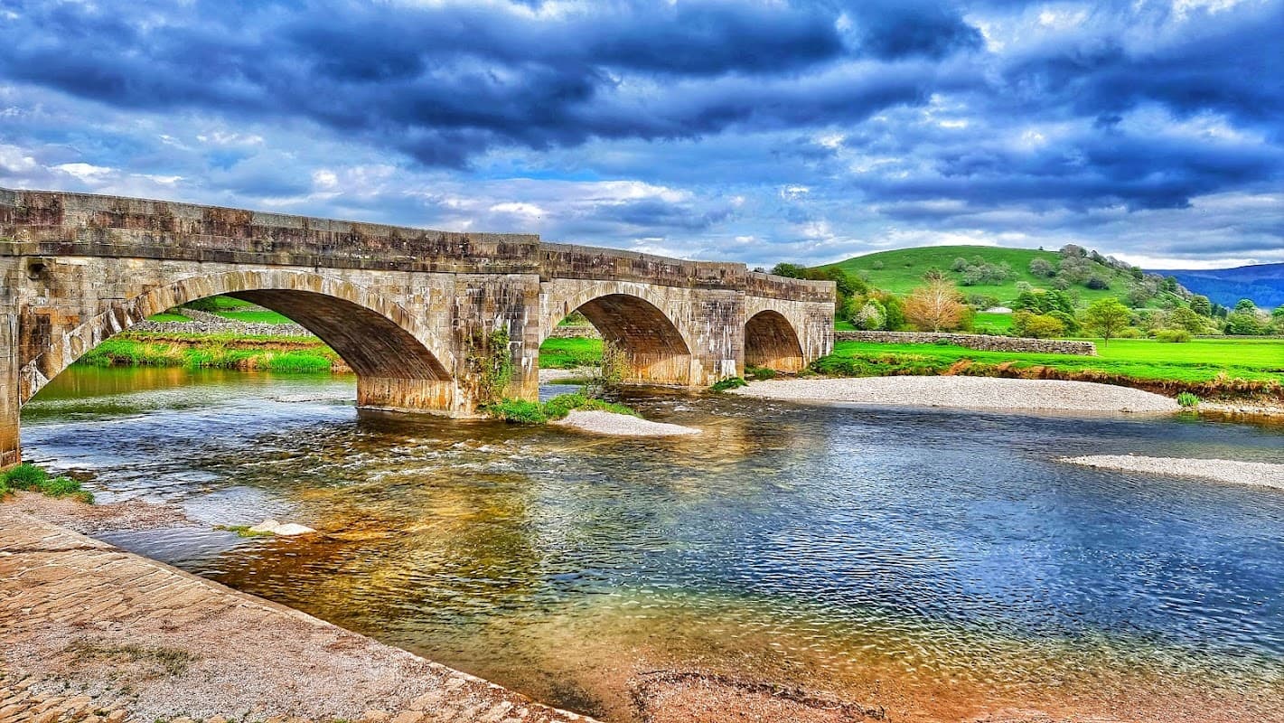 Stone arch bridge over a river, surrounded by lush green fields and dramatic cloudy skies in Appletreewick, Yorkshire.