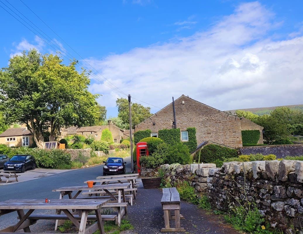 Quaint hotel with stone buildings, picnic tables, a red telephone box, and lush greenery under a blue sky.