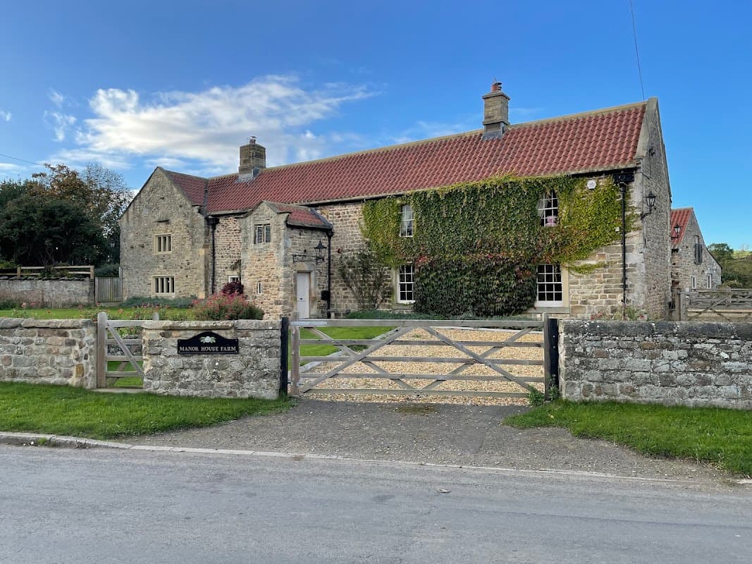 Stone building with a red-tiled roof, surrounded by greenery and a gravel path, featuring a wooden gate.