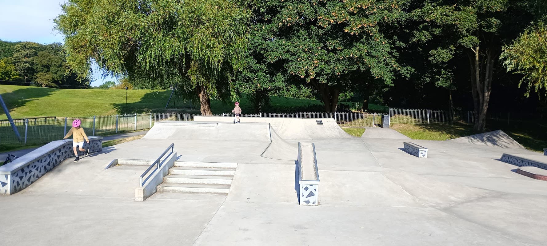 Skatepark with ramps, steps, and a smooth concrete surface, surrounded by trees and green grass.