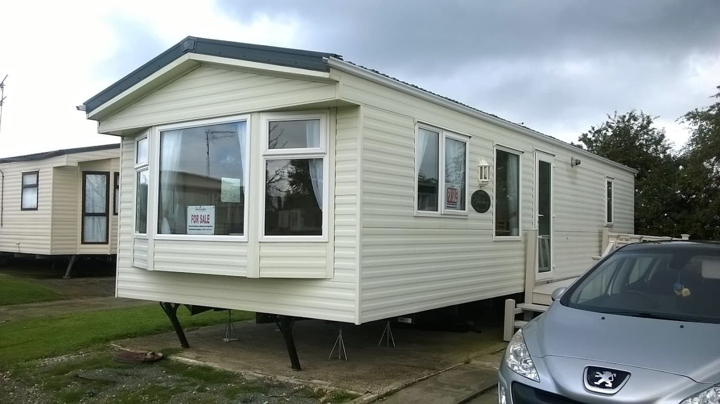 A light-colored mobile home with large windows, parked on grass near a silver car under a cloudy sky.