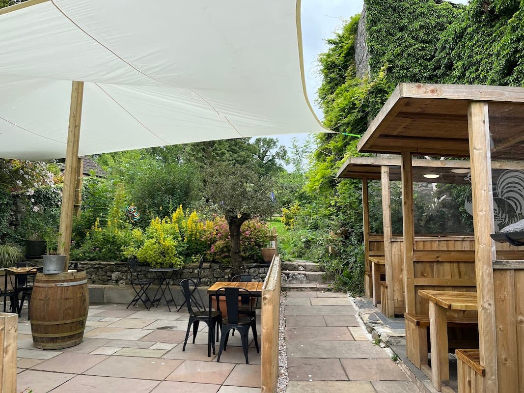 Outdoor dining area with wooden tables, chairs, and a large white canopy, surrounded by lush greenery and flowers.