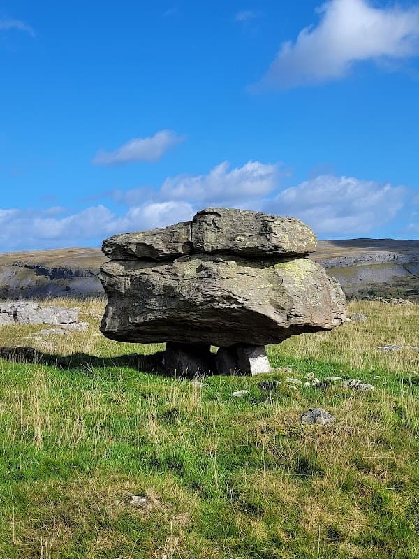 Large, flat boulder balanced on smaller stones, set against a blue sky and grassy landscape in Austwick, Yorkshire.