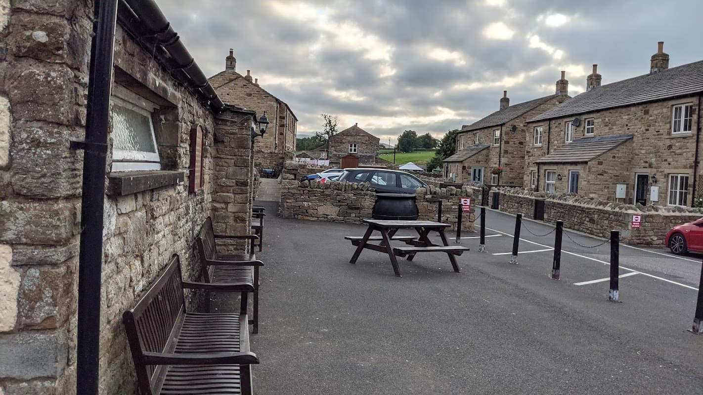 Stone building with benches outside, parking area, and surrounding houses under a cloudy sky in Bainbridge, Yorkshire.