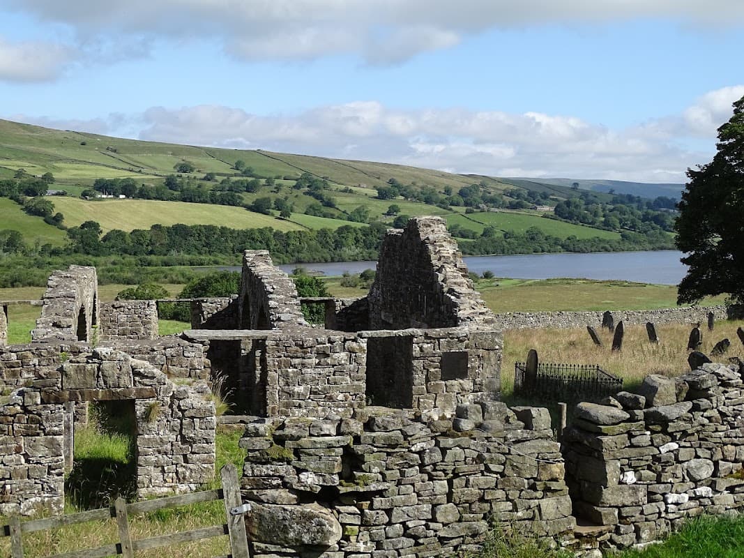 Ruins of stone structures with green hills and Semer Water lake in the background under a partly cloudy sky.