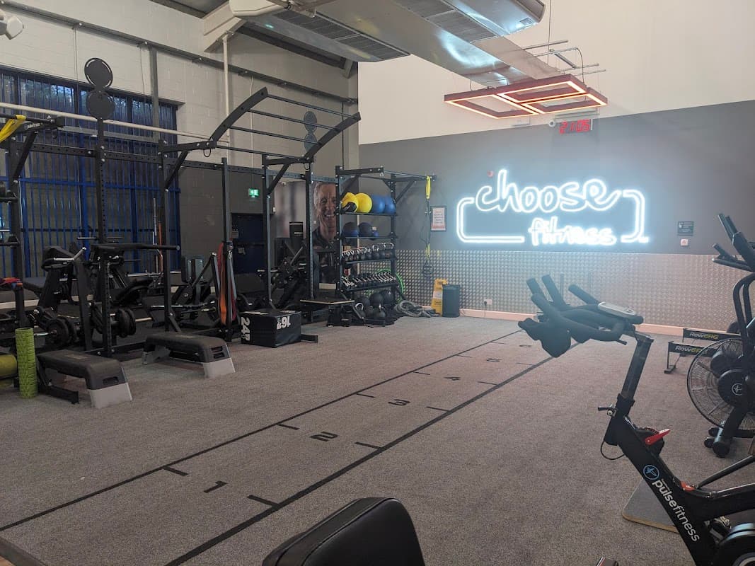 Bright gym interior with workout equipment, weights, and a neon sign reading "choose Fitness."