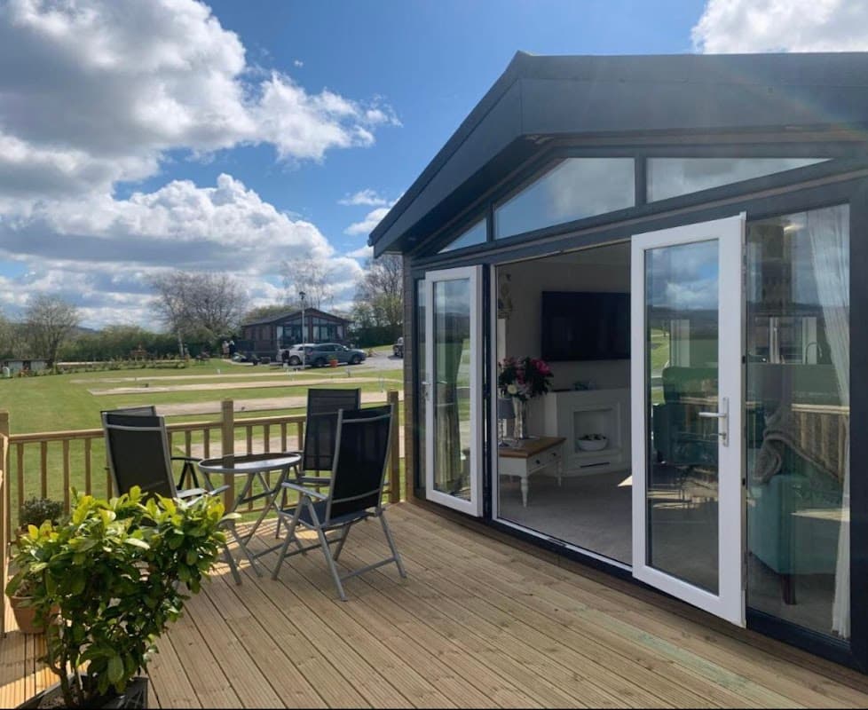 Modern lodge with a deck, outdoor seating, and large windows, set in a green landscape under a blue sky with clouds.