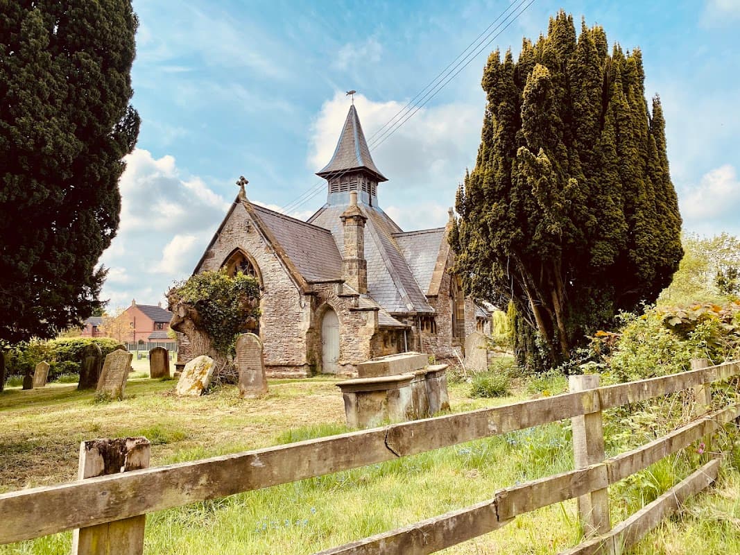 Historic stone church with a tall spire, surrounded by trees and a grassy cemetery, near a wooden fence.