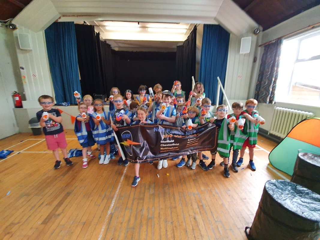 Children holding water guns and a banner inside Bardsey Village Hall, with colorful decorations and a wooden floor.
