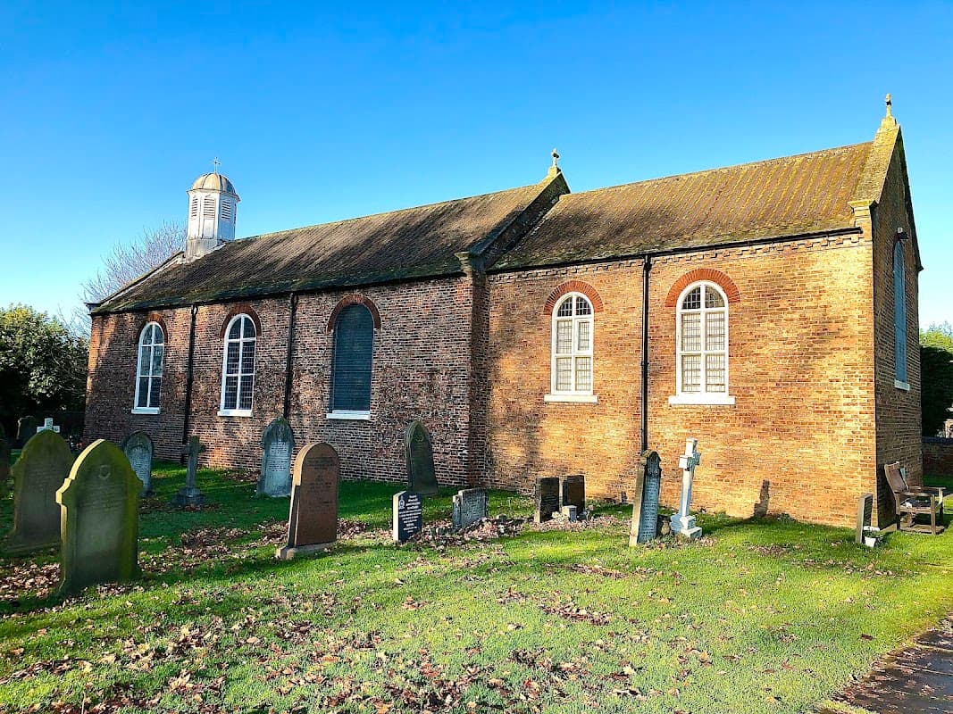 All Saints church in Barlby, Yorkshire, with a brick exterior and gravestones in a grassy cemetery under a clear blue sky.
