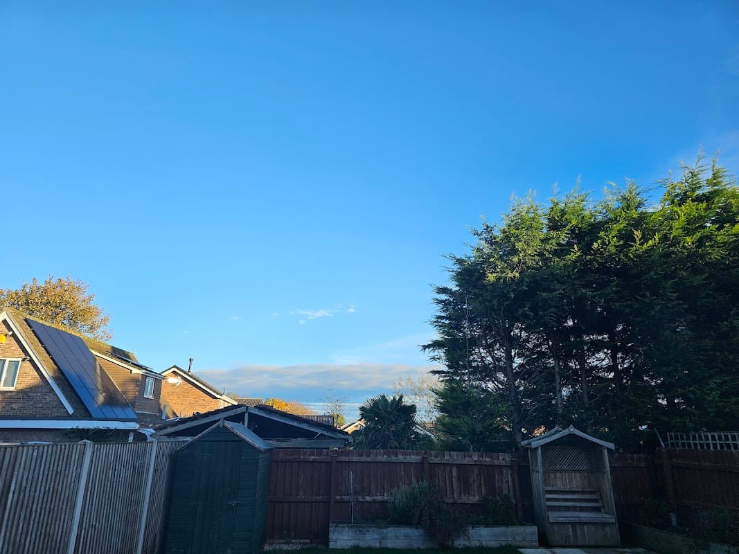 Clear blue sky over residential buildings and greenery, with a wooden fence and garden features in the foreground.