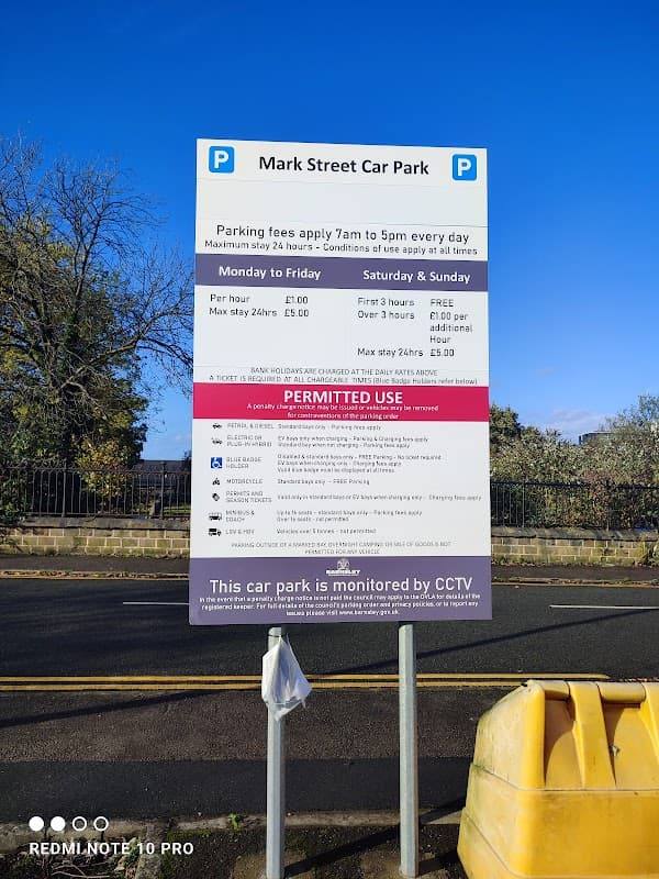 Sign for Mark Street Car Park detailing parking fees, hours, and conditions, with a yellow barrier in the foreground.