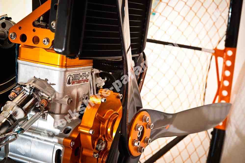 Close-up of a paramotor engine with orange and silver components, showcasing intricate mechanical details and a propeller.