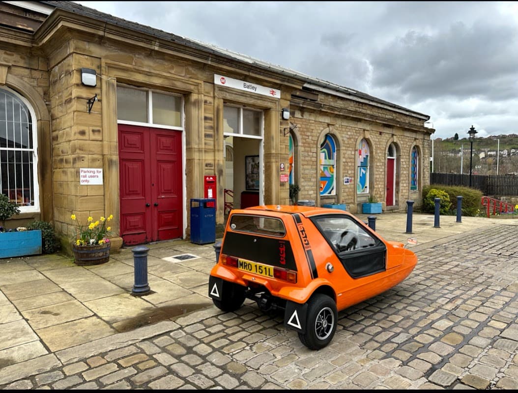 Batley Station features a historic stone building, an orange car parked outside, and colorful planters along the walkway.