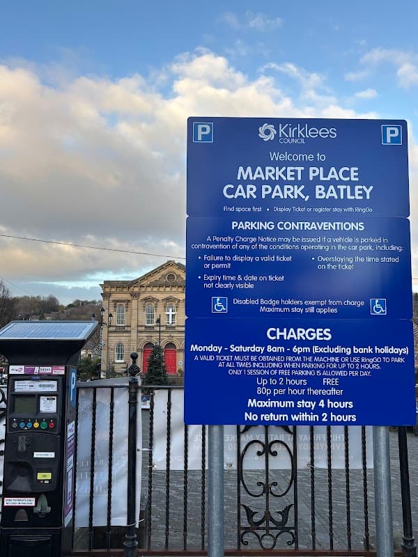 Sign for Market Place Car Park in Batley, displaying parking rules, charges, and payment options. Historic building in background.