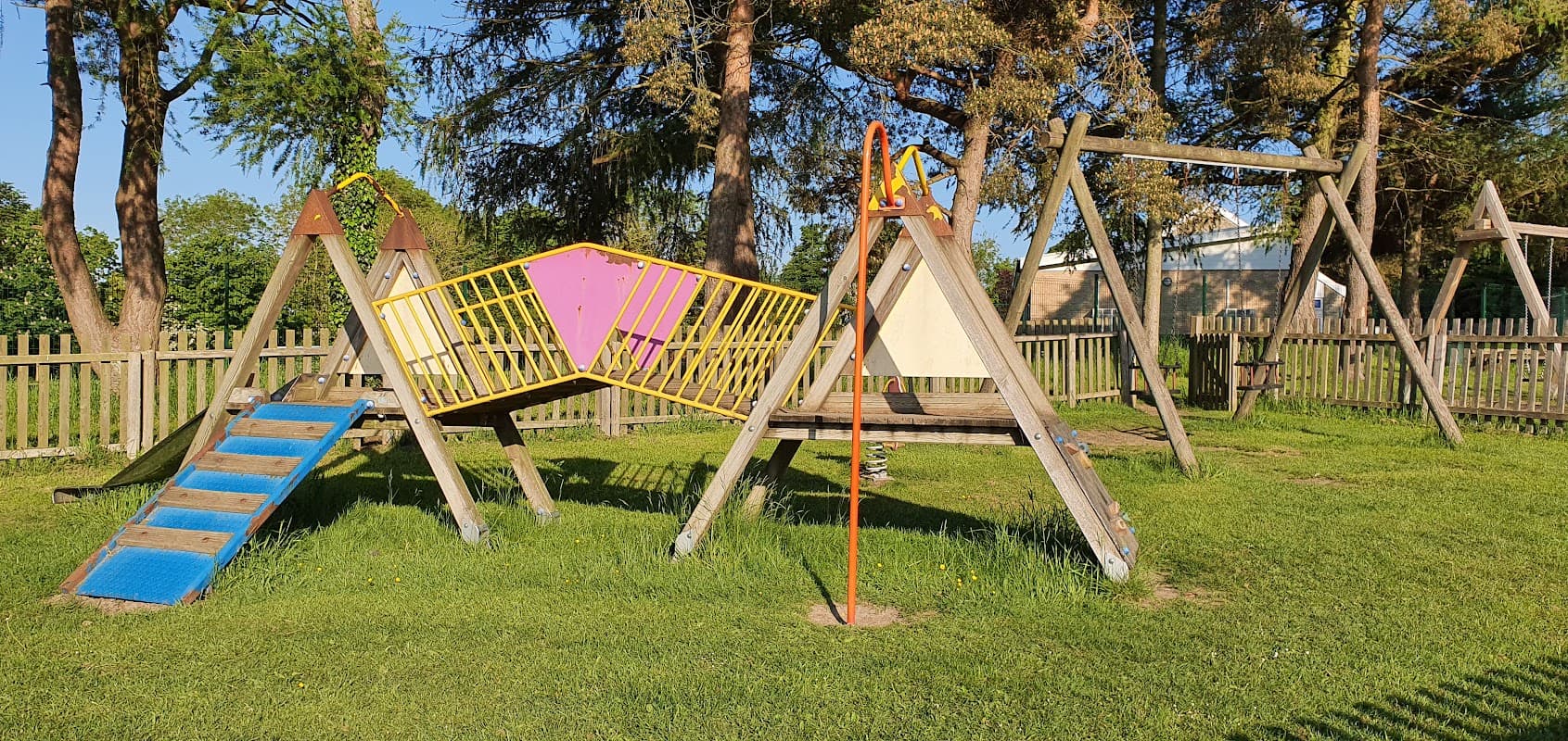 Colorful playground equipment with swings and a climbing frame surrounded by grass and trees in a fenced area.