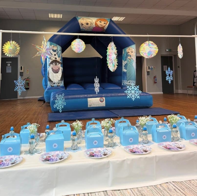 A blue bouncy castle with Frozen characters, decorated tables with blue party boxes and floral arrangements.