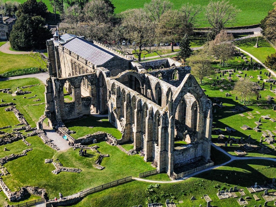 Ruins of Bolton Abbey with arched stone walls, surrounded by green grass and trees, near a cemetery.