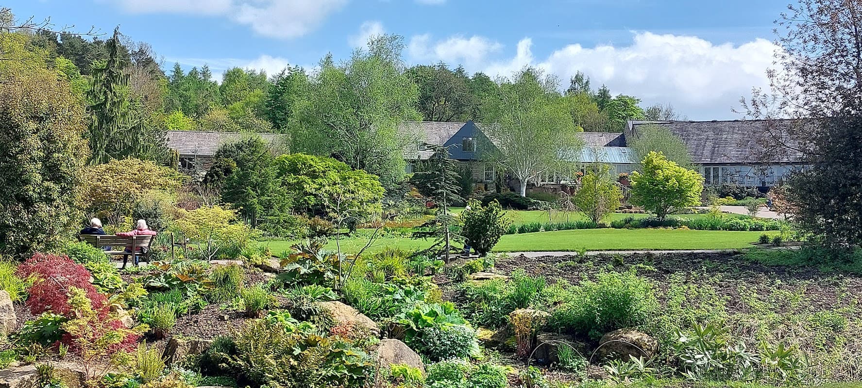 Lush gardens with diverse plants, trees, and seating areas, set against a backdrop of buildings and blue sky.