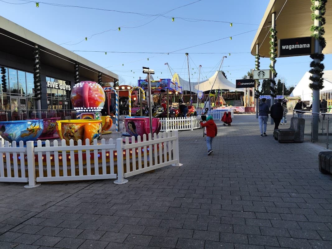 Colorful amusement rides and attractions in a festive car park area, with shoppers walking and string lights above.