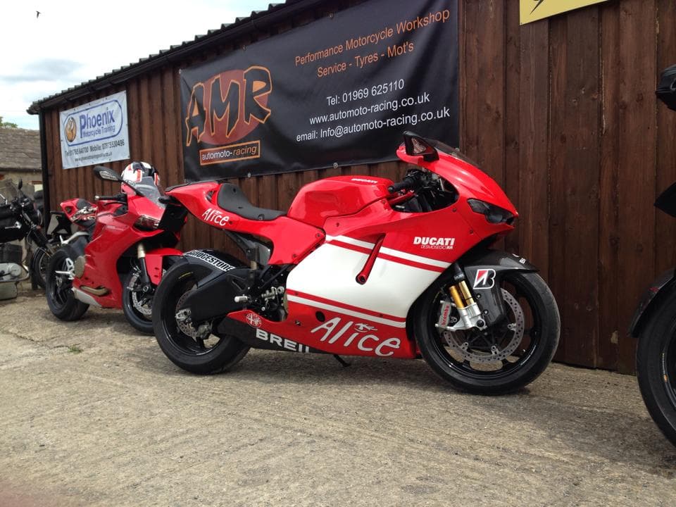 Red Ducati motorcycle with "Alice" branding parked outside a garage, alongside another bike, against a wooden wall.