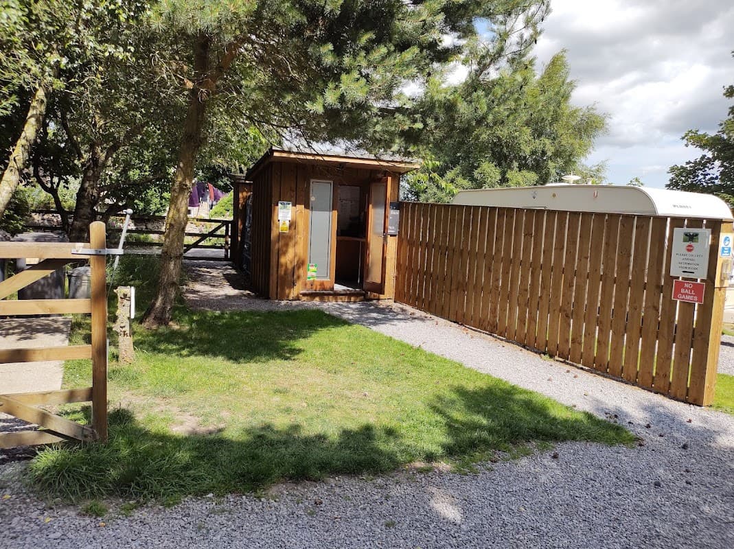Wooden reception cabin with a fenced area, gravel path, and greenery at Cow Close Caravan & Camping in Bellerby, Yorkshire.