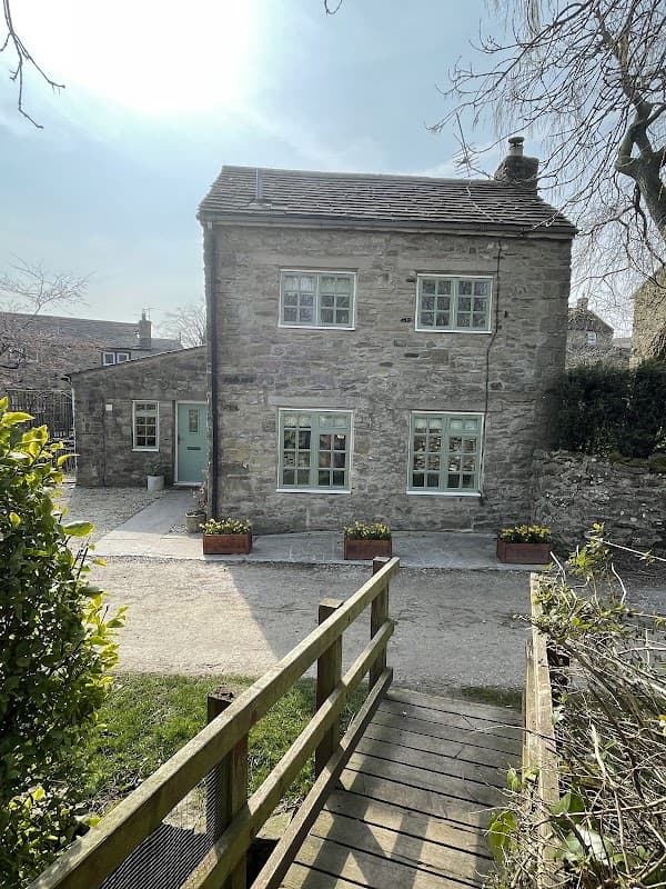 Stone cottage with light blue windows and flower boxes, surrounded by greenery and a wooden walkway.