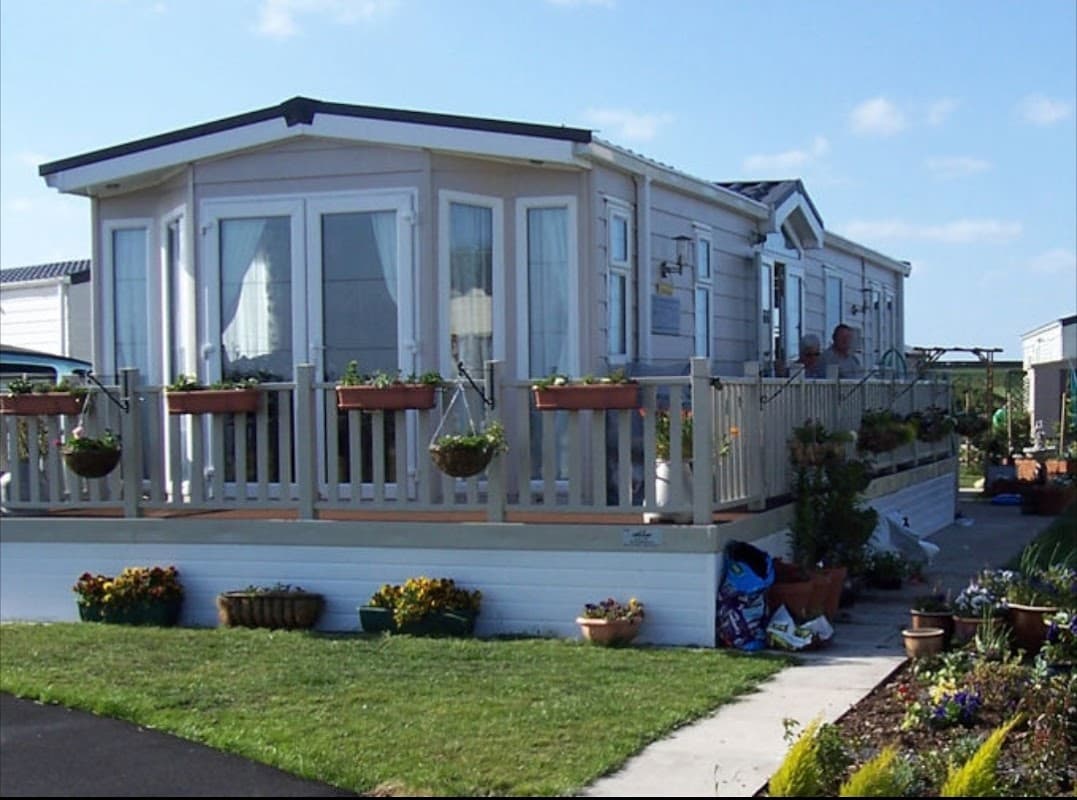 A modern caravan with a deck, surrounded by flower pots and greenery, set against a clear blue sky in Bempton, Yorkshire.