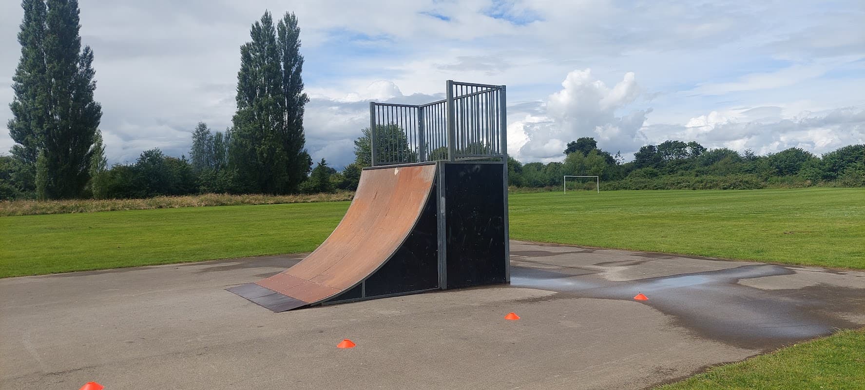 Skate ramp with a metal frame on a concrete surface, surrounded by grass and trees under a cloudy sky.
