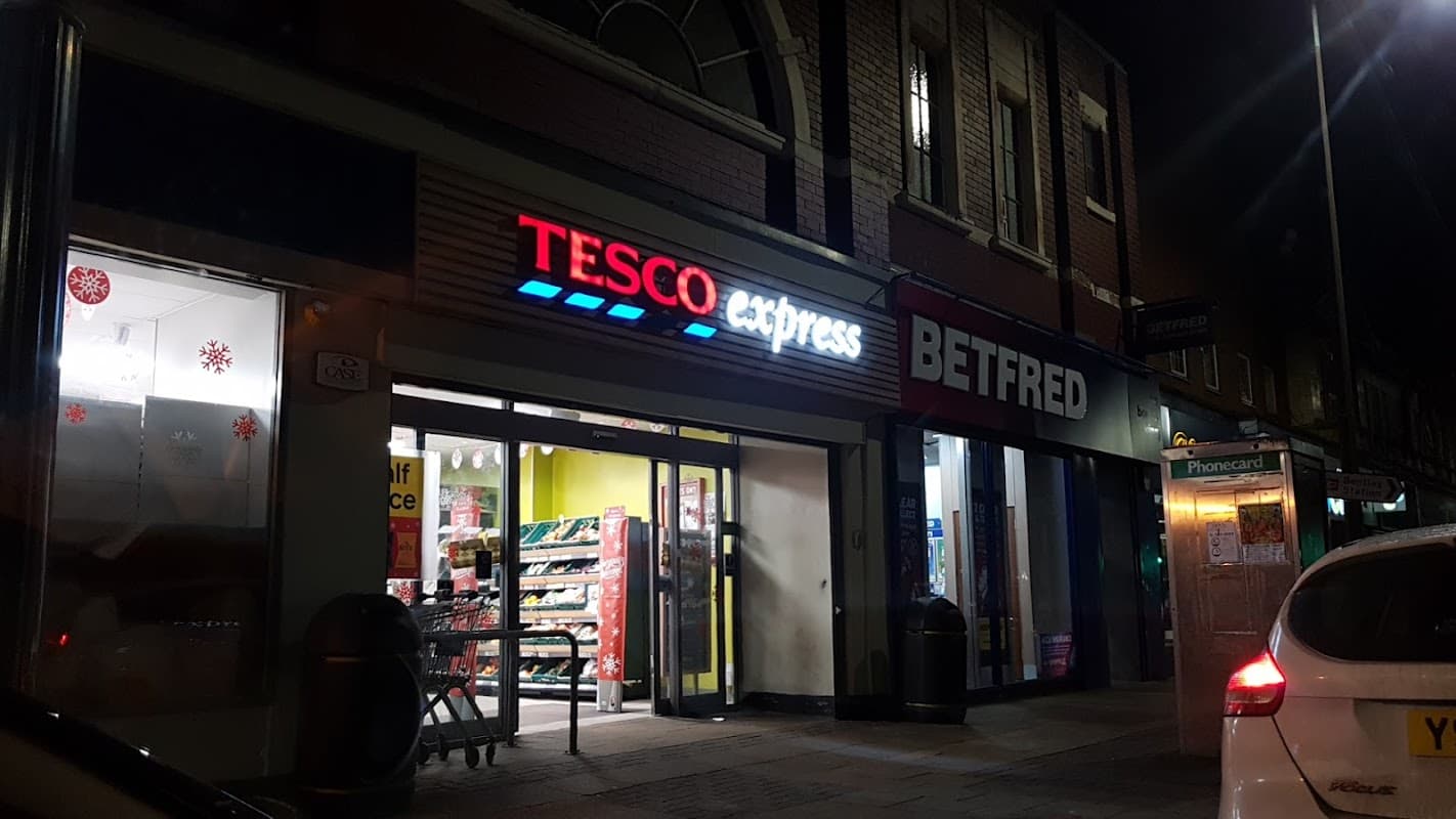 Tesco Express storefront at night, featuring bright signage and nearby Betfred shop, with a parked car in view.