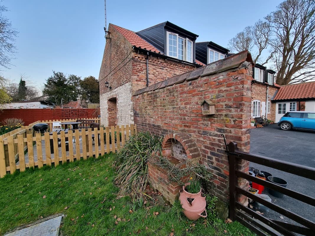 Brick wall with an arch, green grass, and holiday cottages with windows, surrounded by a wooden fence and trees.