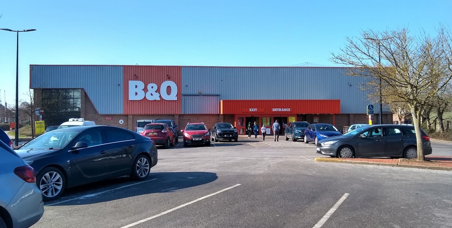 B&Q store exterior with a parking lot filled with cars and shoppers entering the entrance on a clear day.