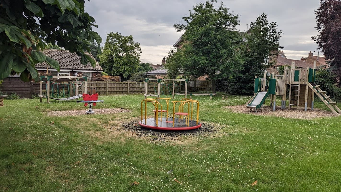 Playground in Bilbrough with slides, swings, a merry-go-round, and grassy areas surrounded by trees and fences.