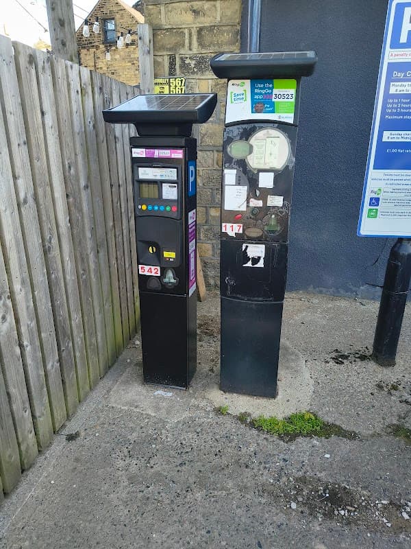 Two parking payment machines next to a wooden fence, with a parking sign visible in the background.