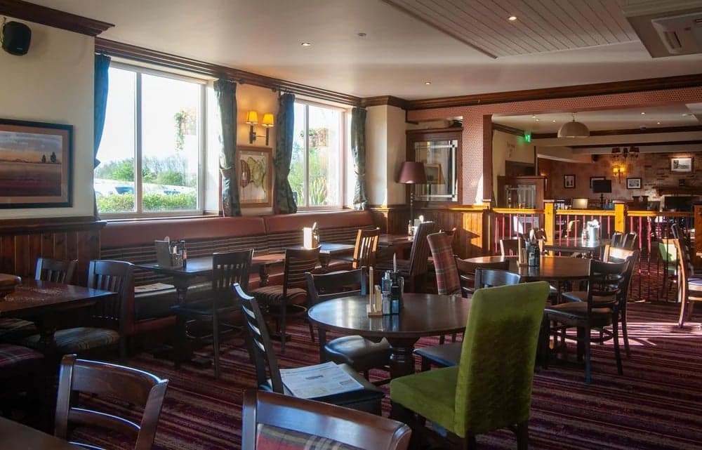 Cozy dining area with wooden tables, chairs, and large windows, featuring plaid decor and warm lighting.