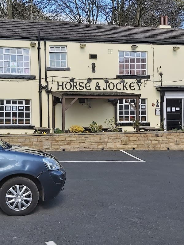 The facade of the Horse & Jockey pub in Birstall, featuring a sign and flower beds, with a parked car in front.
