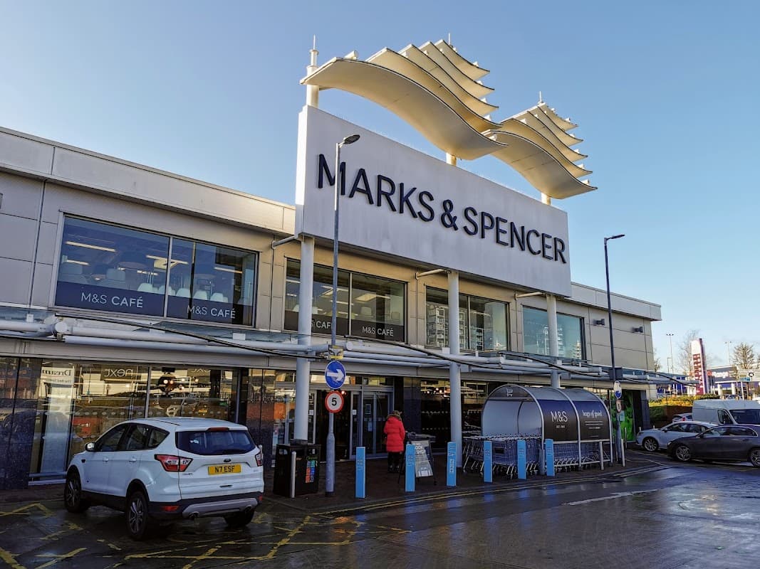 M&S Simply Food storefront with large signage, outdoor seating, and a parked car in front on a sunny day.