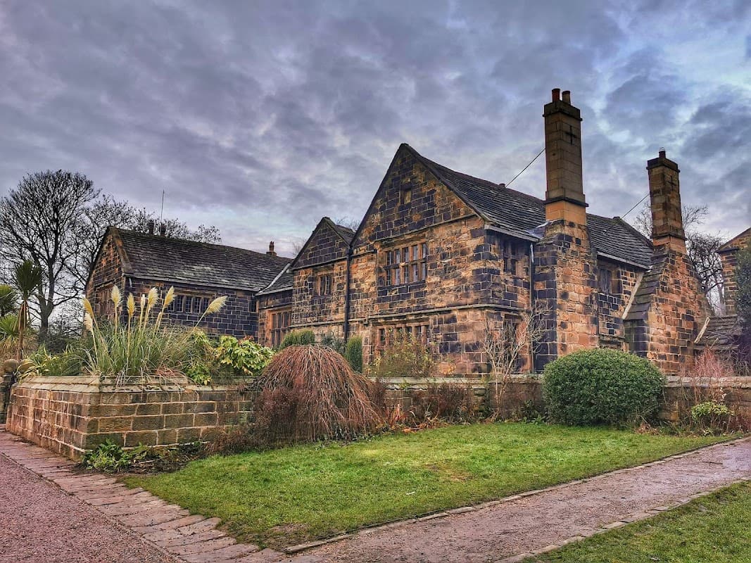 Historic stone building surrounded by lush greenery and garden features under a cloudy sky.