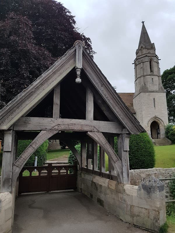 Wooden entrance structure leading to a stone church tower amidst lush greenery and a cloudy sky.