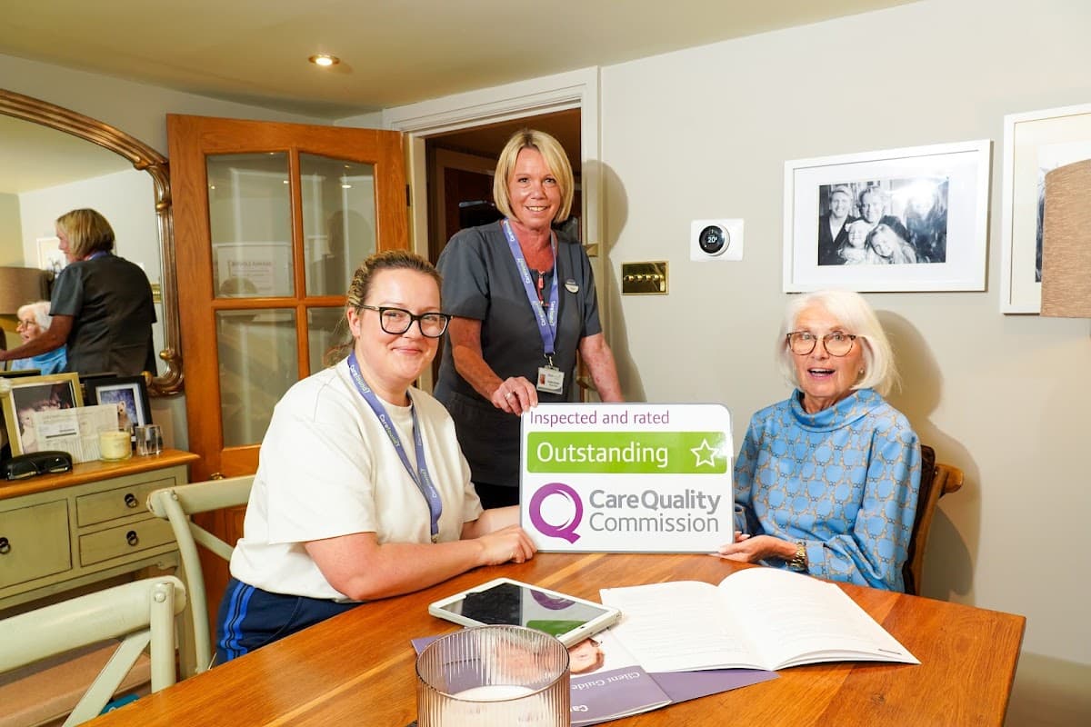 Two caregivers and an elderly woman holding an "Outstanding" Care Quality Commission sign at a cozy home setting.