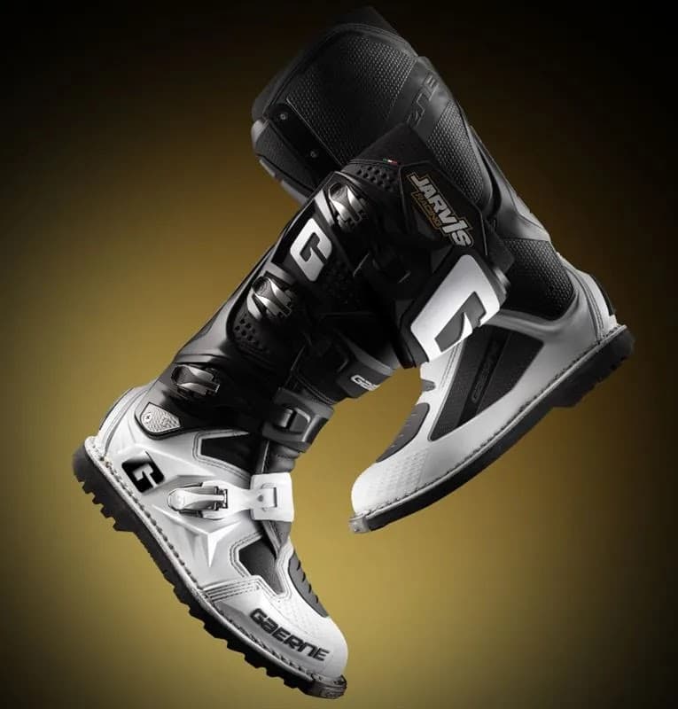 Black and white motocross boots with sleek design, showcasing straps and branding against a gradient background.