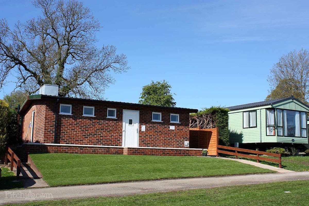 Red brick building with a white door, surrounded by green grass and trees, next to a green caravan.