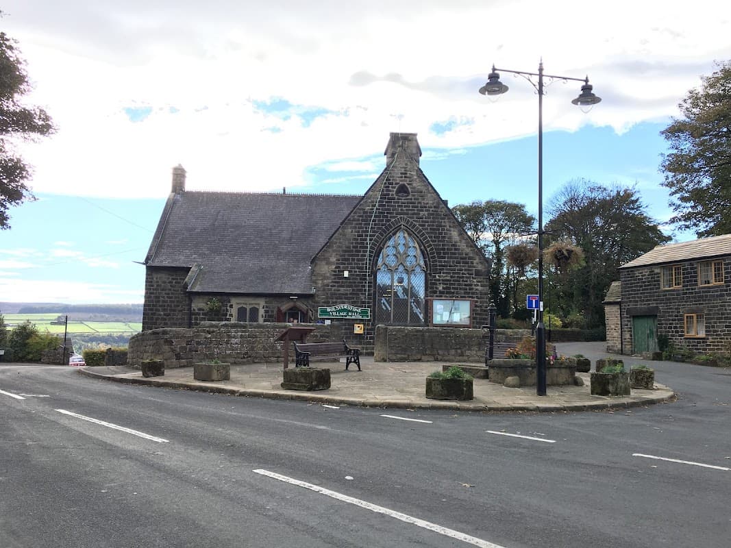 Bolsterstone Village Hall with a large stained glass window, surrounded by greenery and a circular stone seating area.