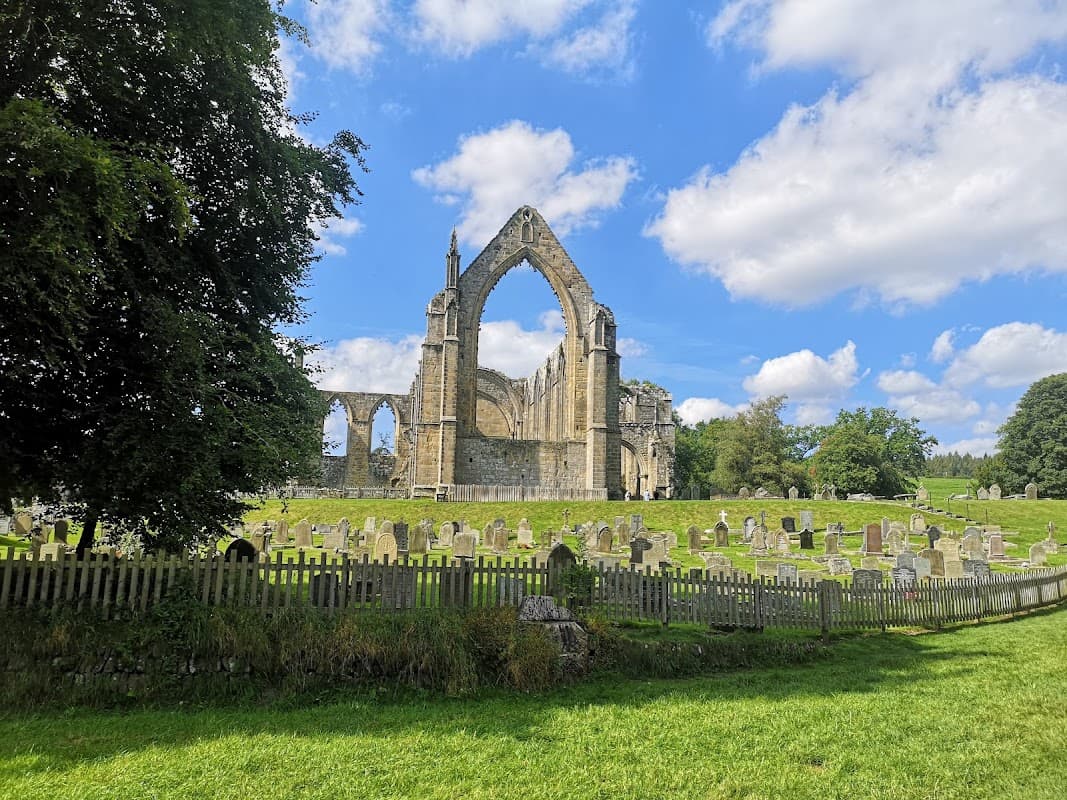 Ruins of Bolton Abbey with a grassy area and gravestones, framed by trees under a blue sky with fluffy clouds.