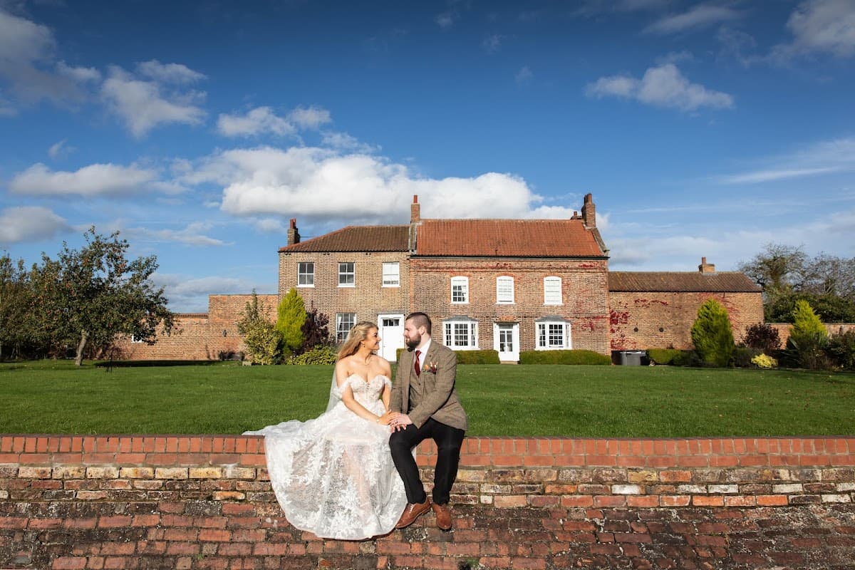 A couple in wedding attire sits on a brick wall in front of Hornington Manor, surrounded by green lawns and blue skies.